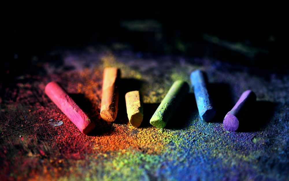 Цветные мелки, разложенные в ряд на темной поверхности, с рассыпавшимся пигментом всех цветов радуги. Символ многообразия, уязвимости и яркости человеческих историй.